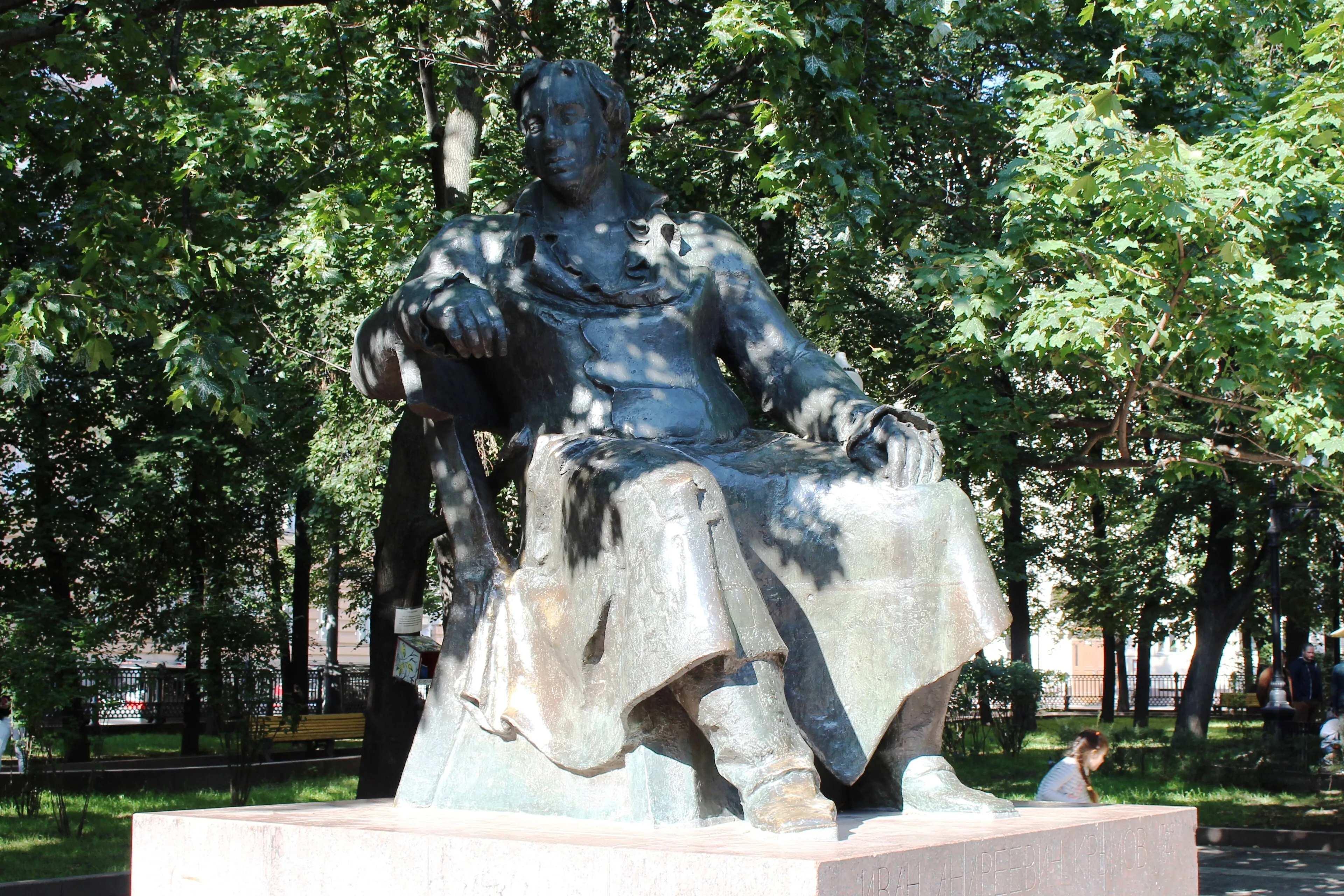 Бронзовый памятник Крылову, сидящему на скамье, среди деревьев